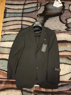 Svart kavaj från Top Tailor - Snygg svart kavaj från Top Tailor med två knappar framtill, klassisk krage och fickor med lock. Tillverkad i stretchigt material för extra komfort och rörelsefrihet. Perfekt för dig som vill ha en stilren och modern look.