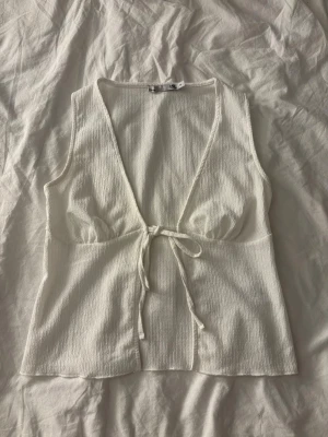 Vit omlottopp från 151® med knyt - Säljer en vit omlottopp från 151® i storlek XS. Toppen är ärmlös, har djup v-ringning och knyt framtill. Tillverkad i ett lätt, strukturerat bomullstyg som känns luftigt och perfekt för varma dagar. Enkel och stilren design som passar till mycket.