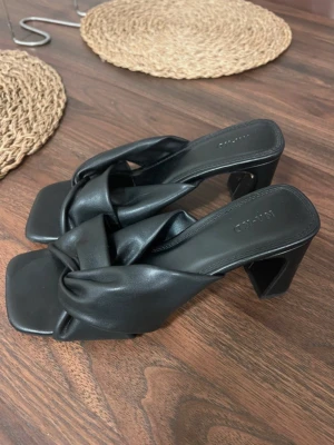 Svarta klackade sandaler från NA-KD - Snygga svarta sandaler från NA-KD med breda, korslagda remmar i konstläder. Skorna har fyrkantig tå och stabil klack som ger en trendig look. Perfekta för dig som vill ha något stilrent men ändå edgy.