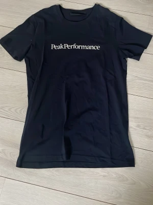 Svart Peak Performance t-shirt - Snygg svart t-shirt från Peak Performance med vit logga tryckt över bröstet. Klassisk rak passform och rund halsringning. Perfekt basplagg till garderoben och enkel att matcha med olika stilar.