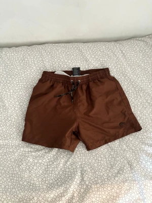 Bruna badbyxor från Lacoste - Snygga bruna badbyxor från Lacoste med klassisk krokodillogo på benet. Modellen har elastisk midja med svart/vit dragsko och sidofickor. Materialet är lätt och snabbtorkande, perfekt för strand eller pool. Helt ny, aldrig använda!