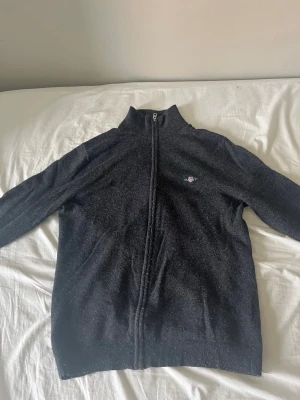 Mörkblå ziptröja med hög krage - Mörkblå ziptröja med hög krage och diskret broderad logga på bröstet. Tröjan har långa ärmar och ribbade muddar vid ärmslut och nederkant. Perfekt för lager på lager och enkel att matcha med olika outfits.