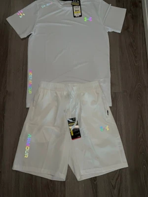 Under Armour set - Säljer ett vitt pyjamasset från Under Armour med t-shirt och shorts. Setet har holografiska loggor och textdetaljer i regnbågsfärger. Materialet är lätt och andas, perfekt för chillkvällar hemma. Elastisk midja på shortsen för extra komfort.