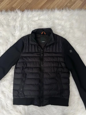 Svart quiltad pufferjacka från Rockandblue - Snygg svart pufferjacka från Rockandblue med quiltad front och släta ärmar. Jackan har dragkedja framtill, två fickor med dragkedja och diskret logga på ärmen. Perfekt för kyliga dagar och enkel att matcha med olika outfits.