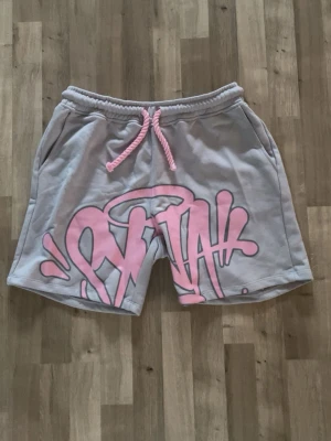 Grå shorts med rosa graffiti och snörning - Säljer ett par grå shorts med rosa graffiti-tryck framtill och matchande rosa snörning i midjan. Shortsen har avslappnad passform, sidofickor och en bakficka med dragkedja. Perfekta för en streetwear-look.