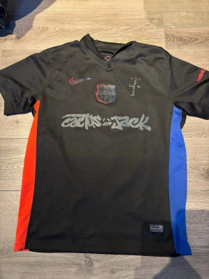 FC Barcelona x Nike Lamine Yamal tröja - Snygg svart FC Barcelona matchtröja från Nike med röda och blå sidopaneler. Tryck med 'Cactus Jack' på framsidan och Lamine Yamal 19 på ryggen. Klubbmärke och Nike-logga på bröstet, UNHCR-tryck bak. Perfekt för dig som älskar fotboll och streetstyle.