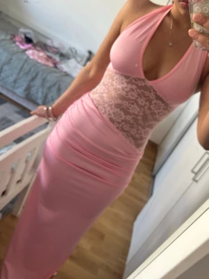 Rosa långklänning med spetsdetaljer - Snygg rosa långklänning med halterneck och djup urringning. Klänningen har spetsdetaljer över magen och ryggen samt knyt i nacken. Figurnära passform och mjukt, stretchigt material som ger en feminin siluett.