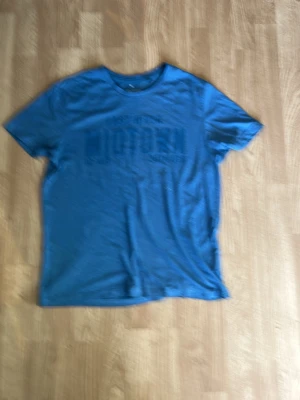 Blå kortärmad t-shirt - En enkel blå t-shirt med rund hals och tryck framtill. T-shirten har korta ärmar och är gjord i mjukt material, perfekt för en avslappnad stil. Passar till jeans eller shorts för en chill look.