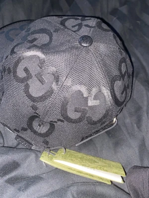Svart keps med GG-mönster från Gucci - Snygg svart keps från Gucci med det klassiska GG-monogrammet i mörkgrått över hela kepsen. Justerbar rem baktill och tillverkad i slitstarkt tyg. Perfekt för dig som vill ha en exklusiv och trendig accessoar.
