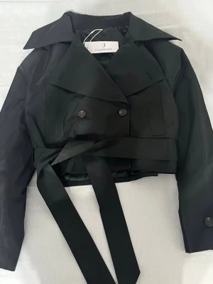 Svart cropped trenchcoat Eva Jonsson - Stilren svart cropped trenchcoat från Eva Jonsson med bred krage, dubbelknäppning och knytskärp i midjan. Jackan har långa ärmar och en modern, kortare passform som ger en edgy look. Perfekt för dig som vill ha en trendig och tidlös jacka.