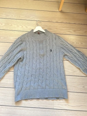 Grå kabelstickad tröja Polo Ralph Lauren - Kabelstickad grå tröja från Polo Ralph Lauren med halv dragkedja och hög krage. Tröjan har ribbade muddar och nederkant samt den klassiska broderade loggan på bröstet. Perfekt för lager-på-lager och chill stil. Har dock ett litet hål på armen, men de går att sy, annars inga problem med de 