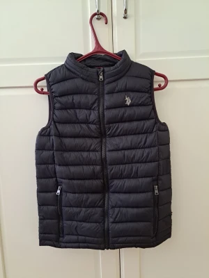 Svart dunväst från U.S. Polo Assn. - Stilren svart dunväst från U.S. Polo Assn. med quiltad design och dragkedja framtill. Västen har två praktiska fickor med dragkedja och en liten logga på bröstet. Perfekt för lager-på-lager och håller dig varm under kyliga dagar.storlek 14-15.