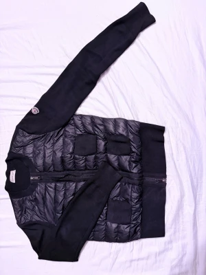 Moncler Cardigan Svart - Svart cardiganjacka från Moncler med quiltad framsida och stickade ärmar. Jackan har dragkedja framtill, ribbade muddar och en Moncler-logga på ena ärmen. Perfekt för lager-på-lager och snygg till jeans. Ni är välkomna att skicka prisförslag ☺️