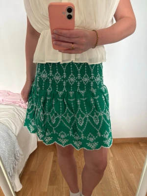 Grön kortkjol med broderade detaljer - Grön kortkjol med vita broderade mönster och vågig kant. Kjolen har smock i midjan för en bekväm passform och är tillverkad i ett lätt tyg som passar perfekt till varmare dagar. Snygg och trendig att matcha med en enkel topp.