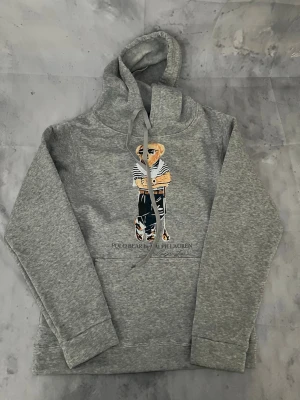 Grå hoodie Polo Ralph Lauren - Snygg grå hoodie från Polo Ralph Lauren med den ikoniska Polo Bear-figuren broderad på bröstet. Hoodien har dragsko i huvan och en klassisk känguruficka framtill. Perfekt för en avslappnad och trendig stil.