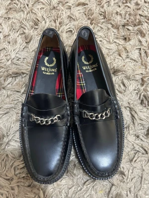Svarta loafers från G.H. Bass Weejuns - Klassiska svarta loafers från G.H. Bass Weejuns med kedjedetalj i silver framtill. Skorna är i blankt läder och har en låg sula. Insidan har ett rutigt foder i rött, svart och gult som ger en snygg kontrast. Perfekta för en stilren och tidlös look.