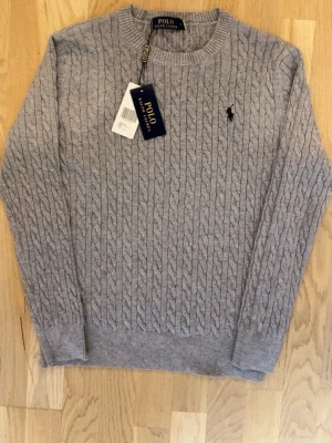 Polo Ralph Lauren Tröja - Kabelstickad grå tröja från Polo Ralph Lauren med rund halsringning och den klassiska broderade loggan på bröstet. Tröjan har långa ärmar och ribbade muddar vid ärmslut och nederkant. Perfekt för en stilren och avslappnad look. Den har använts ett par gånger och har inte taggarna kvar fast är i mycket bra skick.