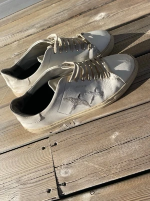 Vita sneakers från Axel Arigato - Snygga vita sneakers från Axel Arigato med fågelmotiv på sidan. Skorna har klassisk låg profil, vita snören och är tillverkade i skinn. Perfekta för dig som gillar stilrena detaljer och streetwear-vibe.