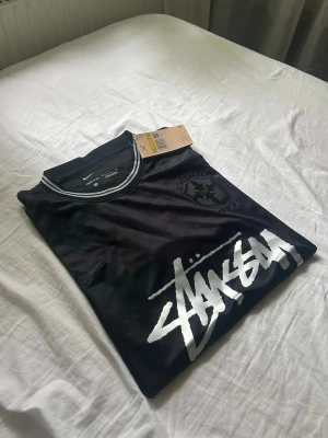 Svart Stüssy x Nike t-shirt - Svart t-shirt från Stüssy och Nike med vit logga framtill och Brasil-märke på bröstet. T-shirten har rund hals med kontrastfärgade detaljer och är tillverkad i ett glansigt material som känns sportigt och fräscht.