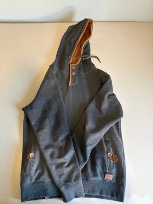 Mörkgrå hoodie med bruna detaljer - Snygg mörkgrå hoodie från Morris med bruna detaljer på insidan av huvan, knappar och armbågslappar. Hoodien har två fickor framtill med knappar och ribbade muddar. Perfekt för en avslappnad och stilren look.