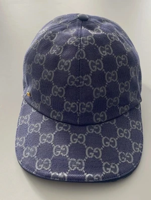 Blå GG-monogram keps från - Säljer en blå keps från  med det klassiska GG-monogrammet i silver över hela kepsen. Kepsen har böjd skärm, justerbart band bak och är tillverkad i canvasmaterial. Perfekt för dig som vill ha en stilren och lyxig accessoar.