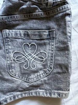 Grå denimshorts från Lindex - PRIS KAN DISKUTERAS! Säljer ett par grå denimshorts från Lindex med snygg broderad detalj på bakfickan. Shortsen har dubbla knappar framtill, bälteshällor och klassiska fickor både fram och bak. Perfekta för sommaren! Knappt använda och köpta för 350-400kr! Storlek 164 men passar som XS. Justerbara i midjan!