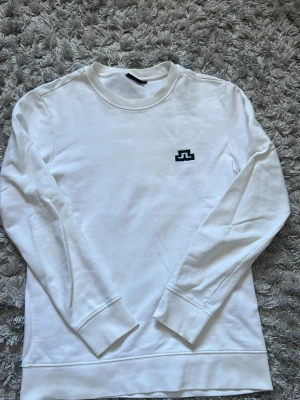 Vit sweatshirt från J.Lindeberg - Vit långärmad sweatshirt från J.Lindeberg med svart logotyp på bröstet. Tröjan har rund halsringning och ribbade muddar vid ärmslut och nederkant. Perfekt för en clean och stilren look.