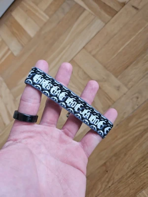 Christian Dior tygarmband med text - Säljer ett svart och vitt tygarmband med broderad text och mönster. Armbandet är gjort av mjukt tygmaterial och har en cool, grafisk stil med kontrasterande färger. Perfekt för dig som gillar unika accessoarer med attityd.