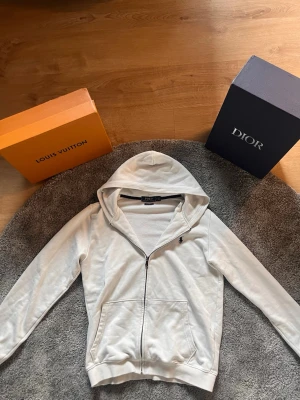 Vit hoodie från Polo Ralph Lauren - Snygg vit hoodie från Polo Ralph Lauren med dragkedja framtill och klassisk huva. Tröjan har två fickor och den ikoniska svarta Polo-loggan broderad på bröstet. Tillverkad i mjukt bomullsmaterial som är skönt att bära. Ask som syns på bilden följer ej med