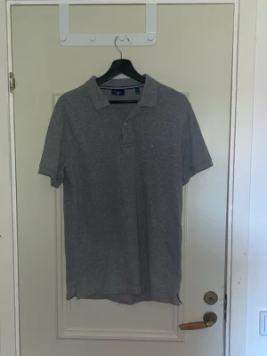 Grå pikétröja från Gant - Klassisk grå pikétröja från Gant med krage och knappar framtill. Tröjan har korta ärmar och liten broderad logga på bröstet. Perfekt för en avslappnad men ändå stilren look.