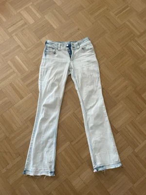 skitsnygga vita bootcut jeans - Säljer ett par vita bootcut jeans med ljusblå slitningar och råa, fransiga benslut. Jeansen har dubbla knappar i midjan, klassiska fem fickor och en cool kontrastdetalj vid gylfen. Perfekta för dig som gillar en avslappnad och trendig stil.