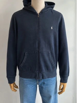 Mörkblå hoodie från Polo Ralph Lauren - Mörkblå hoodie med dragkedja från Polo Ralph Lauren. Klassisk design med vit broderad logga på bröstet, två fickor framtill och huva. Tillverkad i mjukt bomullsmaterial, perfekt för en avslappnad och stilren look.