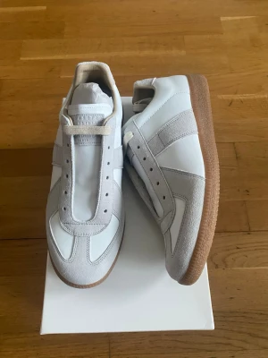 Vita sneakers med beige detaljer - Stilrena vita sneakers från Maison Margiela med beige mockadetaljer och klassisk gummisula. Skorna har en låg silhuett och är tillverkade i en mix av läder och mocka. Perfekta för dig som gillar minimalistisk och tidlös design.