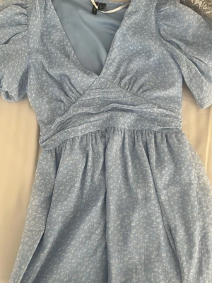 Ljusblå omlottklänning Vero Moda XS - Supersöt ljusblå omlottklänning från Vero Moda med kort ärm och diskret mönster. Klänningen har v-ringning, markerad midja och volangdetaljer. Perfekt för sommarens alla tillfällen!