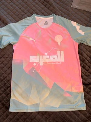 Marocko fotbollströja - Säljer en unik fotbollströja från Puma i pastellfärger med rosa och ljusblå toner. Tröjan har tryck på bröstet med arabisk text och emblem, samt geometriska mönster. Tillverkad i lätt och snabbtorkande material, perfekt för träning eller match.
