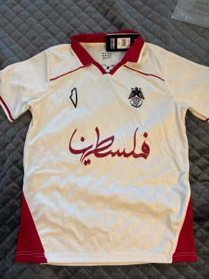 Vit och röd fotbollströja Paliroots - Vit fotbollströja från Paliroots med röda detaljer och krage. Broderad logga med svart fågel och fotboll på bröstet, samt röd arabisk text framtill. Tillverkad i lätt och ventilerande material, perfekt för träning eller match.