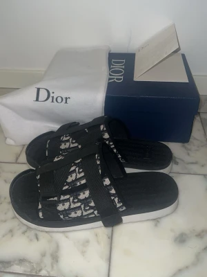 Dior tofflor - Säljer ett par svart och vita  sandaler från Dior med klassiskt Dior-mönster på remmarna. 🔥Sandalerna har breda textilband och justerbara spännen för skön passform. Väldigt bra skick, inga skador och ser riktigt fräscha ut ✈️ skriv om du har frågor 👍