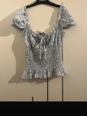Blommig blå volangblus med knyt - Supersöt blommig blå blus med volangdetaljer och knyt framtill. Smockad midja och rynkad nederkant ger en feminin siluett. Korta puffärmar och v-ringning. Perfekt för en somrig look!