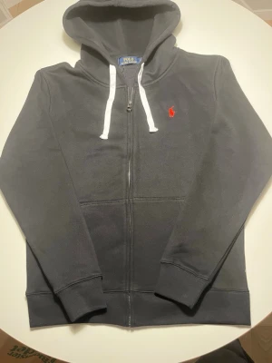 Svart hoodie från Polo Ralph Lauren - Säljer en svart hoodie från Polo Ralph Lauren med vit dragsko och röd broderad logga på bröstet. Hoodien har hel dragkedja, två fickor fram och ribbade muddar. Perfekt för dig som gillar klassisk och stilren design.