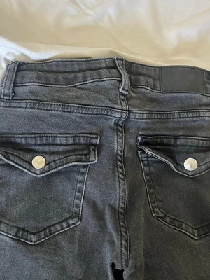 Svarta jeans från 157 med fickdetaljer - Säljer ett par svarta jeans från 157 med snygga ficklock och silverfärgade knappar bak. Jeansen har klassisk femficksdesign och är gjorda i stretchigt denimtyg för bekväm passform. Perfekta till en avslappnad streetstil. (lite slitna nertill)