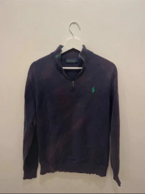 Mörkblå halvzip från Polo Ralph Lauren - Mörkblå stickad tröja från Polo Ralph Lauren med halv dragkedja och grönt broderat logotyp på bröstet. Tröjan har lång ärm och ribbade muddar. Perfekt för en stilren och avslappnad look.