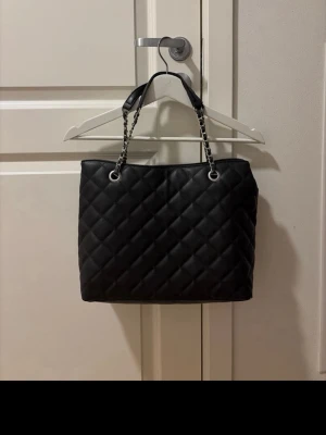 Svart quiltad handväska med kedjerem - Snygg svart handväska i quiltat mönster med kedjeremmar i silverfärg och inslag av läder. Väskan har en klassisk, rektangulär form och är tillverkad i konstläder. Perfekt för dig som gillar stilrena accessoarer med en lyxig touch.
