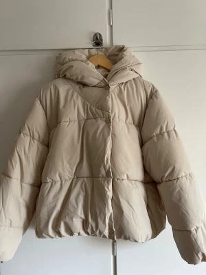 Beige pufferjacka från C&A - Stilren beige pufferjacka från C&A med huva och breda kanaler. Jackan har en oversized passform och stängs med tryckknappar framtill. Perfekt för kalla vinterdagar och enkel att matcha med olika outfits. -stor luva