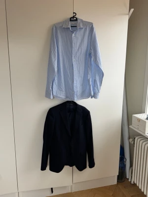 Kostympaket från Riley  - Klassisk skjorta från these glory days  i ljusblått med tunna vita ränder. Skjortan har lång ärm, knappar framtill och en snygg krage. Tillverkad i bomull för en skön och luftig känsla. Perfekt för dig som gillar stilrena och tidlösa plagg. Kostymen är storlek 46 med en grymt snygg passform. Nypris 3500 kr för kostymen och 799 för skjortan. Perfekt för studenten eller andra trevliga tillställningar 