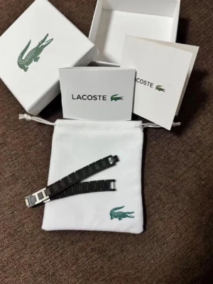 Svart armband från Lacoste  - Stilrent svart armband från Lacoste i metall med rektangulära länkar och graverad krokodil-logga. Armbandet har en modern design med diskreta mönster på länkarna och passar perfekt för dig som gillar minimalistiska accessoarer.