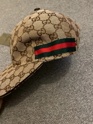 Gucci beige keps med GG-mönster - Snygg beige keps från Gucci med klassiskt GG-monogram över hela kepsen. På sidorna finns de ikoniska gröna och röda ränderna. Kepsen är tillverkad i canvasmaterial och har en böjd skärm. Perfekt för dig som gillar lyxiga accessoarer och streetstyle.