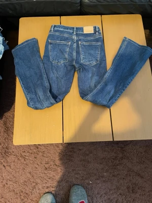 Blå bootcut jeans med läderpatch - Säljer ett par klassiska blå bootcut jeans med två bakfickor och läderpatch på linningen. Jeansen har en tidlös tvätt och är tillverkade i denim med snygga sömdetaljer. Perfekt för dig som gillar en avslappnad och trendig look.