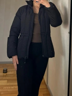 EDC by Esprit jacka - Svart jacka från EDC by Esprit. Jackan har en dragkedja framtill och två fickor med dragkedja i midjehöjd. Kragen är ribbad och insidan av jackan är fodrad med ett mönster av EDC-logotypen. Jackan har även en liten röd etikett med EDC-logotypen längst ner på vänster sida.