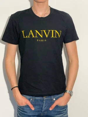Svart Lanvin t-shirt med gul logga - Säljer en svart t-shirt från Lanvin med stor gul logotyp på bröstet. T-shirten har rund hals och korta ärmar, perfekt för dig som gillar stilrena designer och vill sticka ut med ett exklusivt märke.