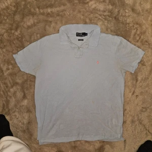 Ljusblå piké från Polo Ralph Lauren - Ljusblå pikétröja från Polo Ralph Lauren med klassisk krage, knappslå och korta ärmar. Tröjan har en liten rosa broderad logga på bröstet och är tillverkad i mjuk bomull för en bekväm känsla. Size taggen stämmer inte och sitter mer som en stor S eller lite mindre M